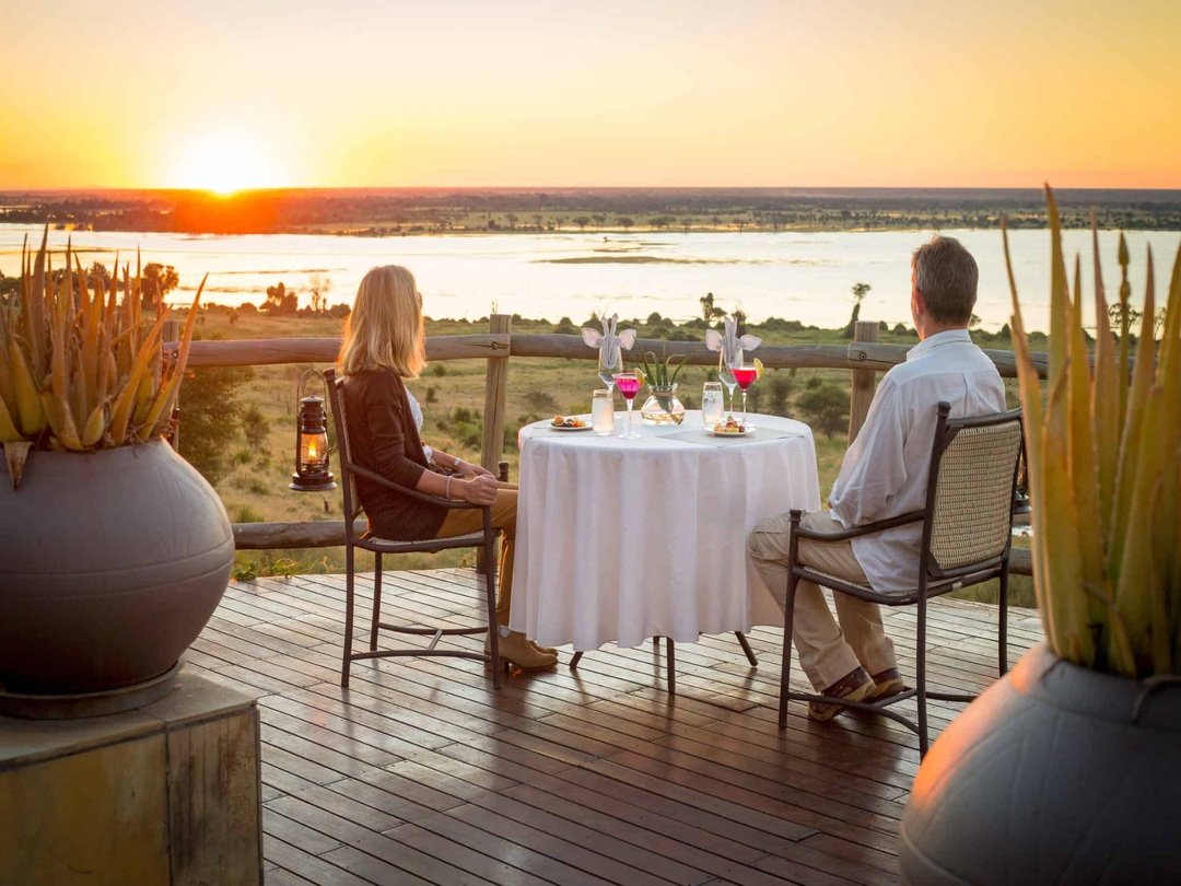 A couple at the banks of the Zambezi River enjoys a breathtaking sunset as they wait for a romantic dinner experience. Surrounded by the peaceful beauty of nature, the golden evening light creates a magical atmosphere where they can relax, connect, and celebrate their love while creating unforgettable honeymoon memories in the heart of Africa.
