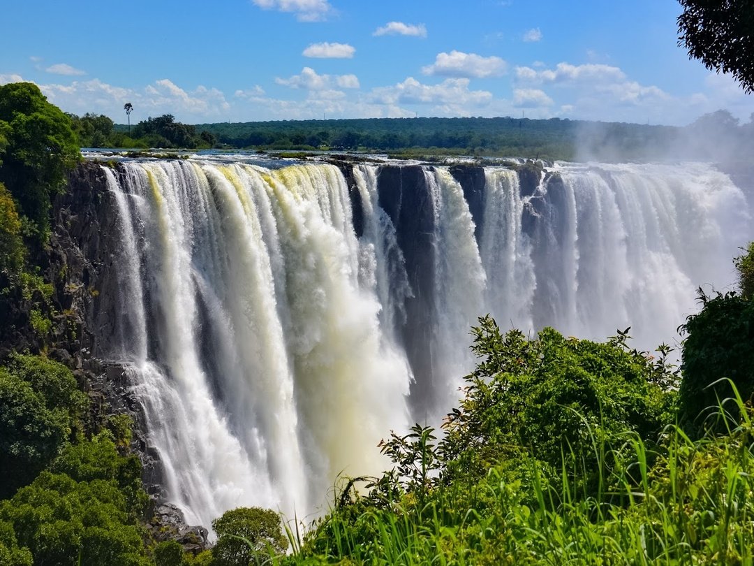 The Victoria Falls cascading dramatically over the Zambezi River, surrounded by lush green vegetation, mist, and breathtaking natural scenery in Southern Africa.