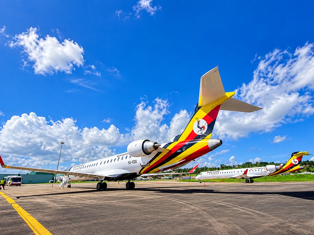 Two Uganda Airlines passenger aircraft parked on an airport apron beneath a bright blue sky with white clouds.